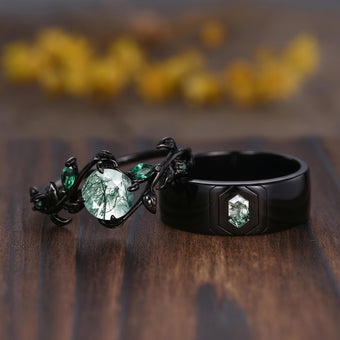 Two black rings displayed on a wooden surface with a blurred yellow floral background. One ring features a large oval clear stone with a green tree-in-water design, and the other is a matte black ring with a smaller hexagonal green stone.