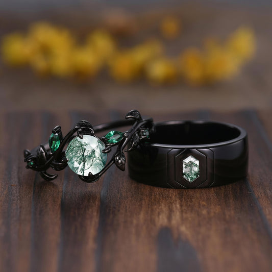 Two black rings displayed on a wooden surface with a blurred yellow floral background. One ring features a large oval clear stone with a green tree-in-water design, and the other is a matte black ring with a smaller hexagonal green stone.